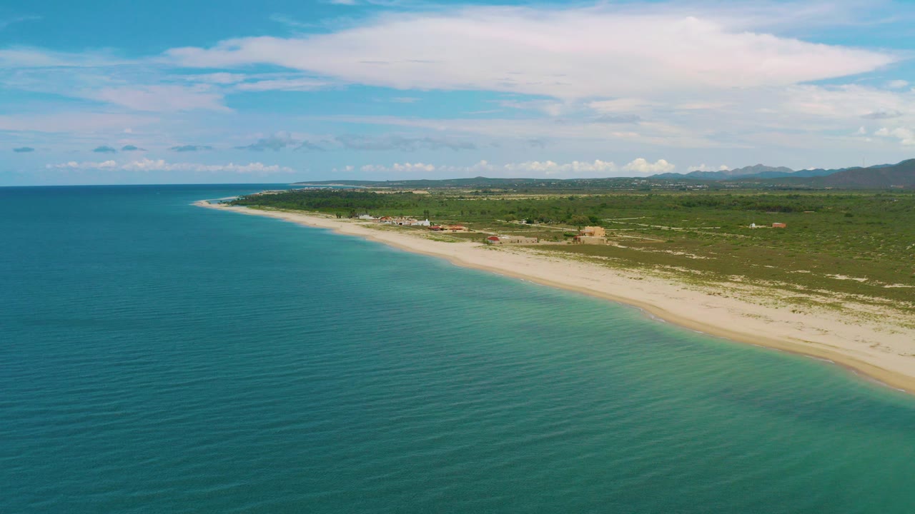 Opening aerial frame over La Ribera, Baja California Sur, with teal sea filling the foreground, a pale beach curving into the distance, green vegetation inland, distant mountains, bright clouds, and no visible people.