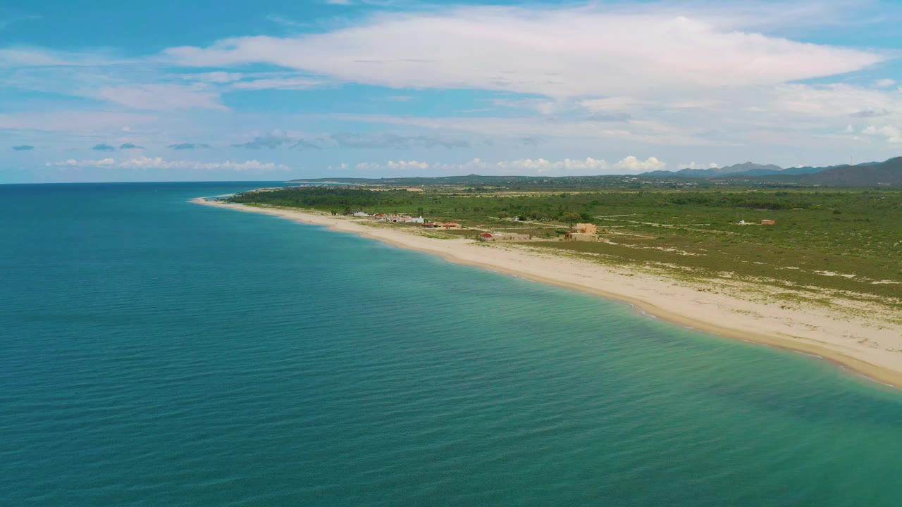 Clear sea water and mild waves run along a wide beach with low greenery inland.