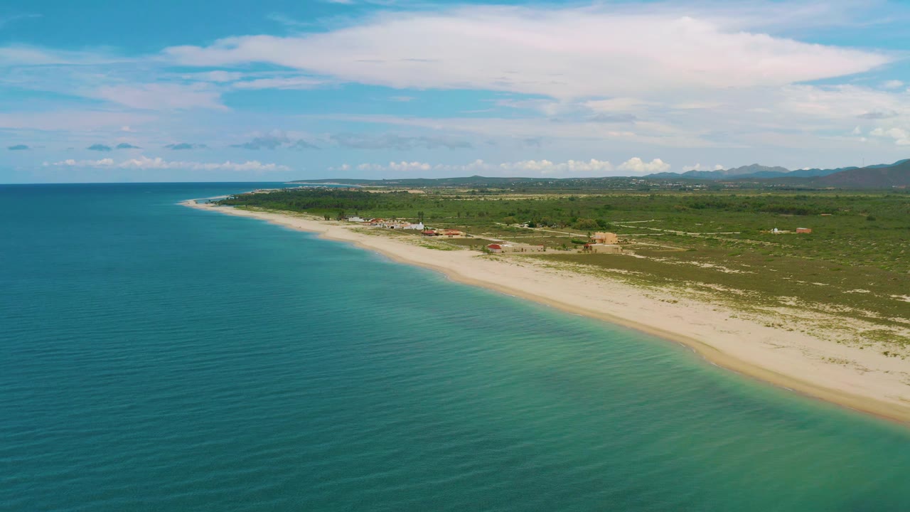 Wide aerial frame over La Ribera, Baja California Sur, Mexico, with calm blue-green water across the foreground, a narrow sand strip on the right, low scrubland, scattered buildings, distant hills, and no visible people.