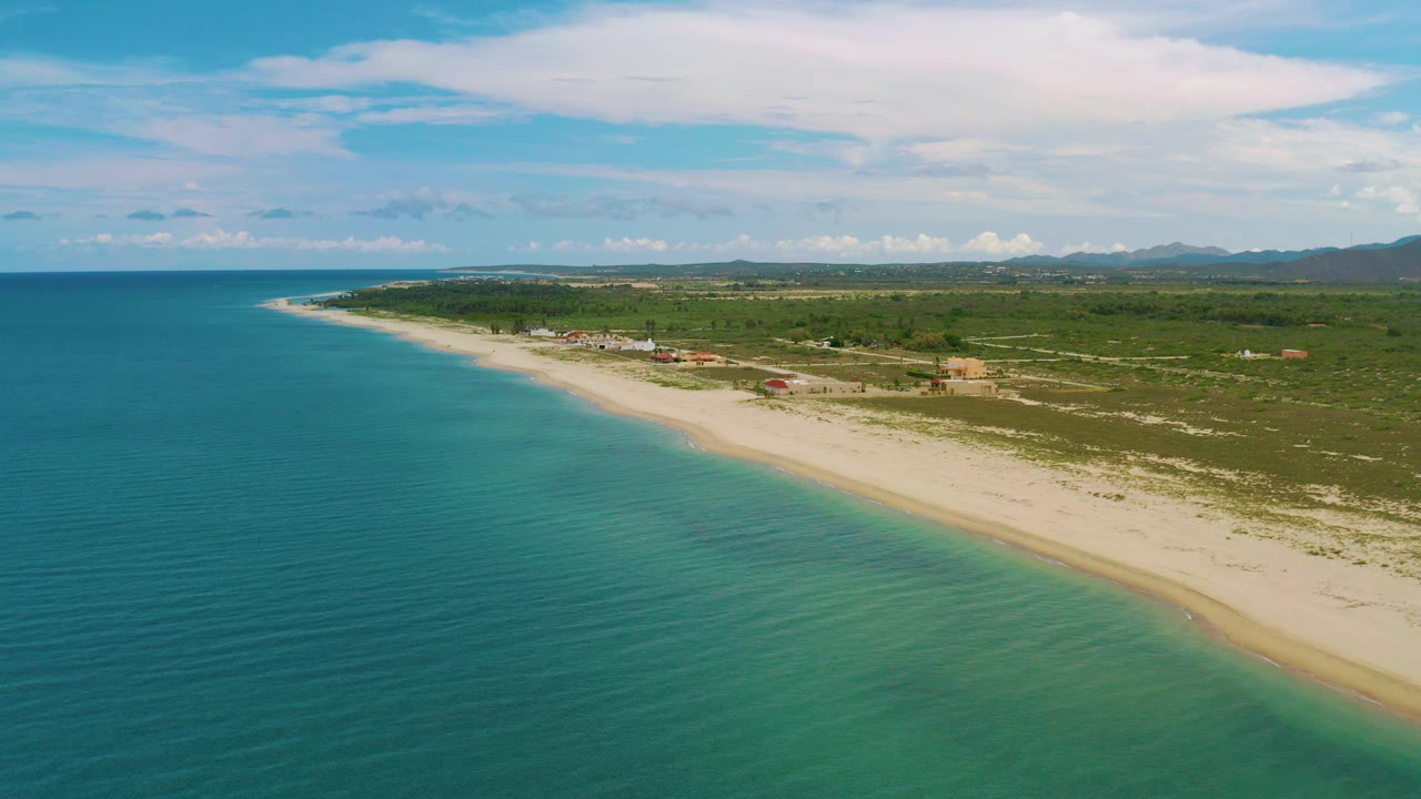 Aerial view over La Ribera, Baja California Sur, with turquoise sea at left, a pale beach curving inland, low green vegetation, distant mountains, and no visible people.