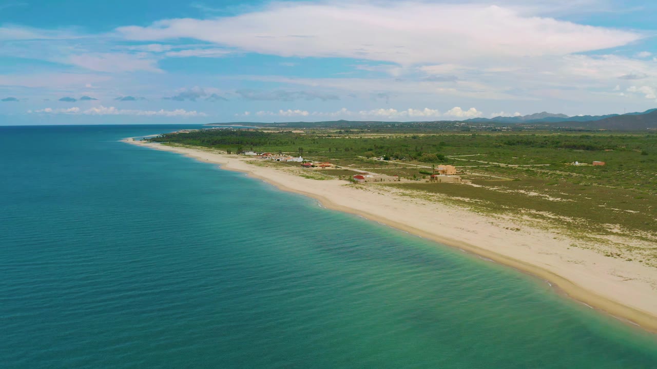Aerial frame of La Ribera shoreline showing blue-green water in the foreground, a narrow surf line, pale sand, scattered inland buildings, distant hills, and no visible people.