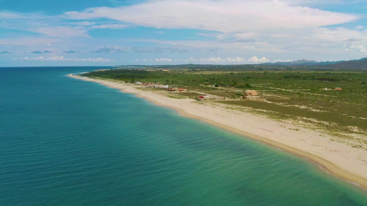 Drone-like view of open sea, pale shoreline, low vegetation, and distant mountains.