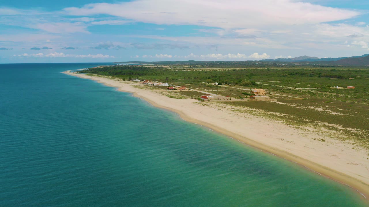 Drone view over La Ribera, Baja California Sur with turquoise water on the left, a pale beach curving into the distance, inland scrub, scattered buildings, distant hills, and no visible people.