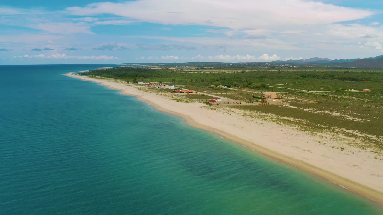 La Ribera aerial view with calm turquoise water, a wide strip of light sand, open scrubland, beachside buildings, a far mountain line, layered clouds, and no visible people.