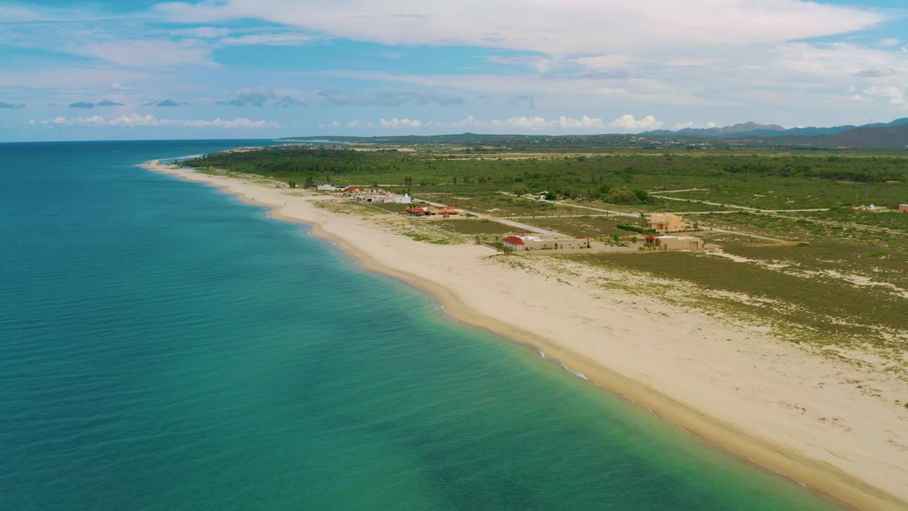 Opening aerial frame over La Ribera, Baja California Sur, with blue ocean at left, a pale sandy beach curving into the distance, low inland vegetation, scattered buildings, distant mountains, and no visible people.
