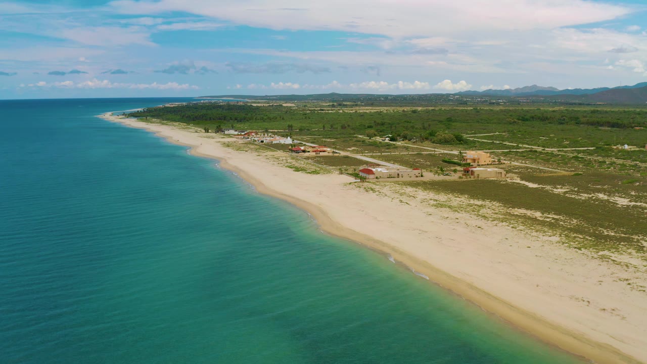 La Ribera aerial frame with calm blue-green ocean, pale beach sweeping from foreground to distance, inland scrubland with scattered structures, mountain ridges on the horizon, and no visible people.