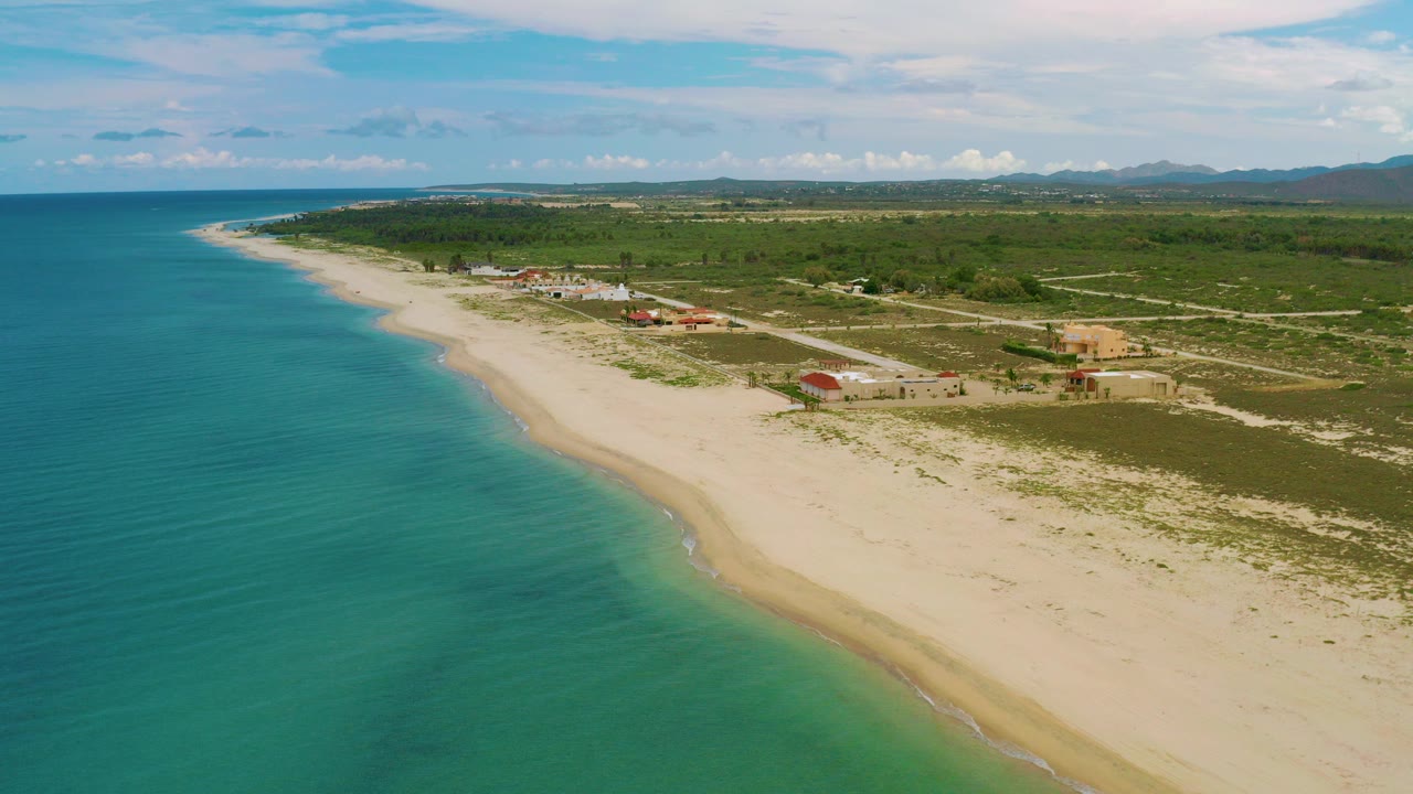 Aerial view over La Ribera, Baja California Sur, with turquoise sea on the left, a pale beach curving inland, scattered coastal buildings, distant hills, and no visible people.