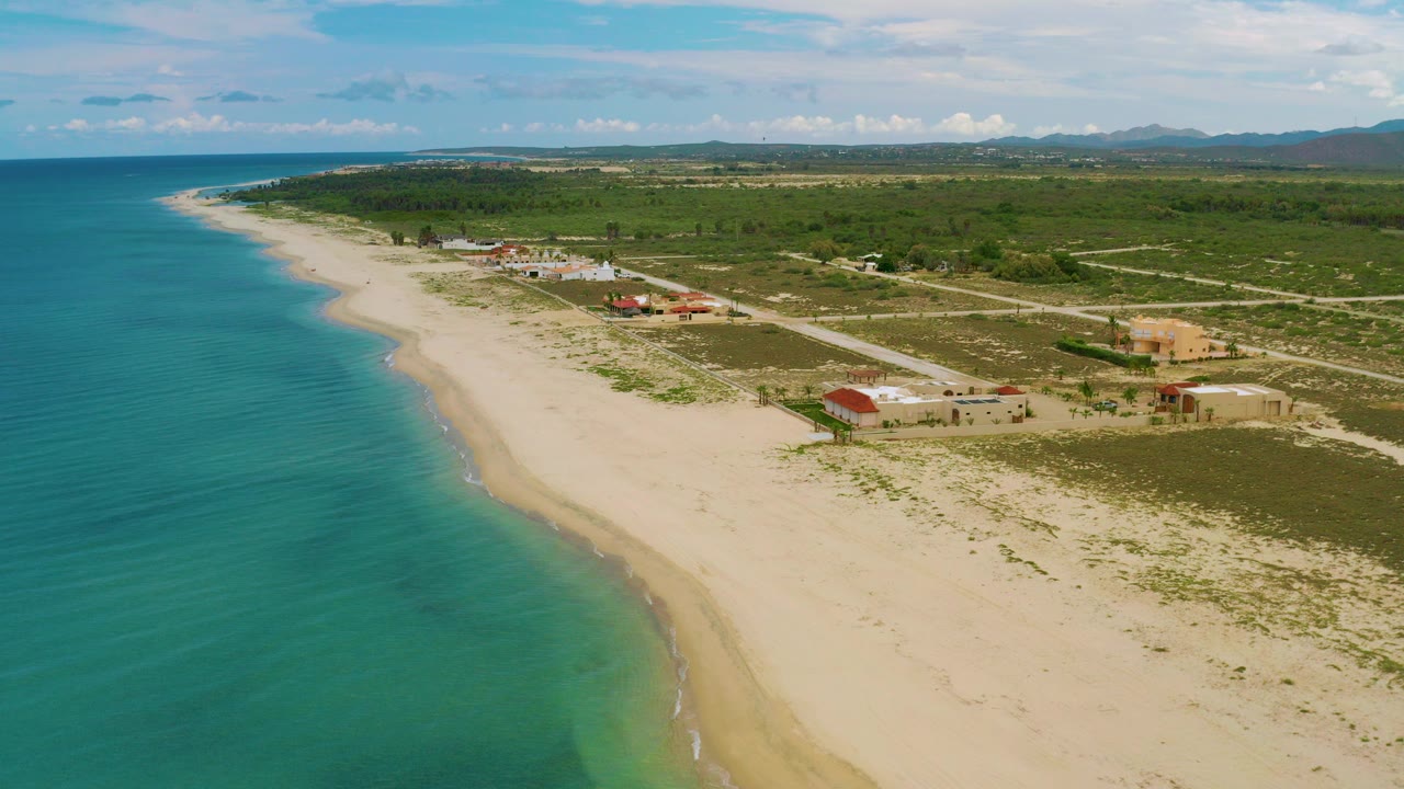 High aerial view over La Ribera, Baja California Sur, with turquoise water on the left, a pale beach curving past low buildings and scrubland, and no visible people.