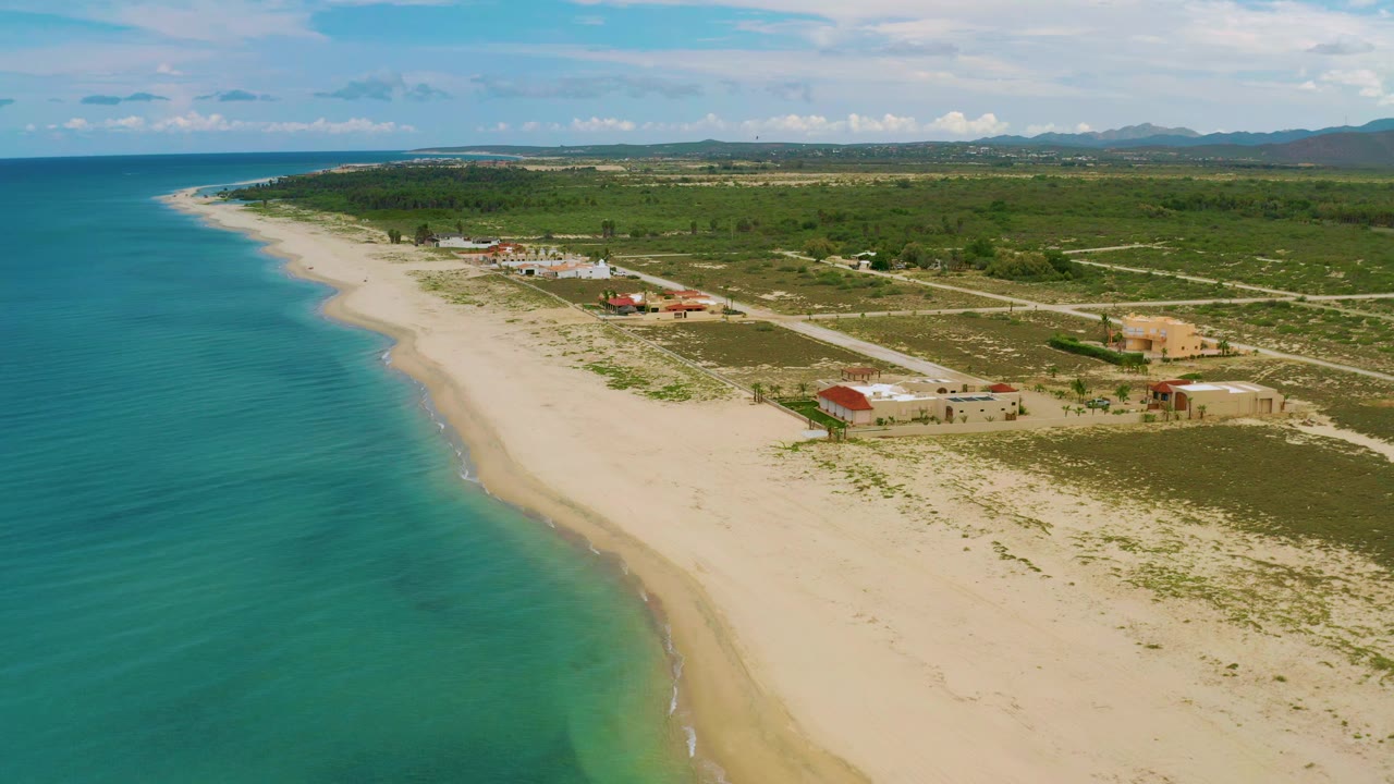 Aerial frame over La Ribera showing blue-green ocean, a broad sandy shoreline, several low buildings near roads, scrub-covered land, distant mountains, and no visible people.