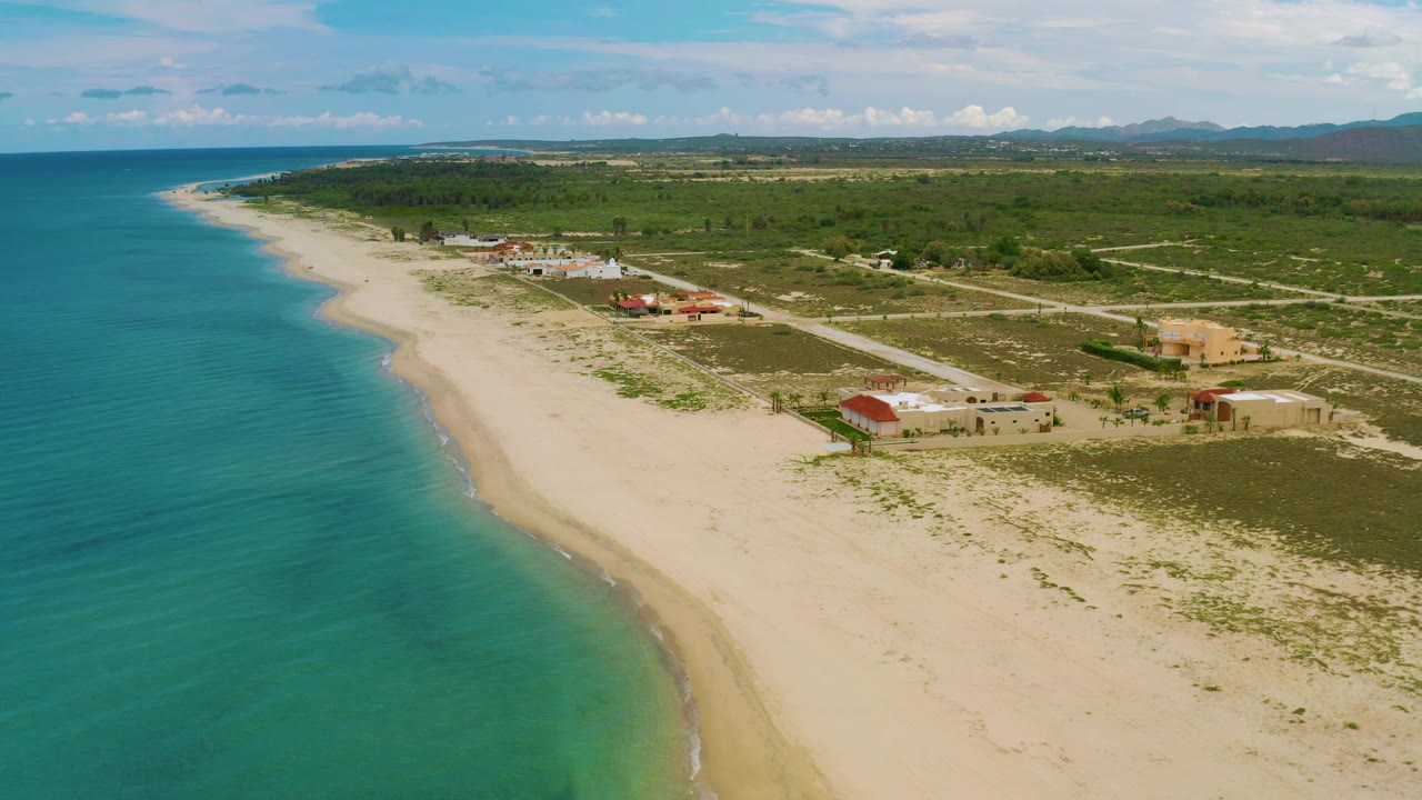 La Ribera aerial frame with the turquoise sea along the left edge, pale sand beach below, rural roads and low structures inland, green scrubland beyond, and no visible people.
