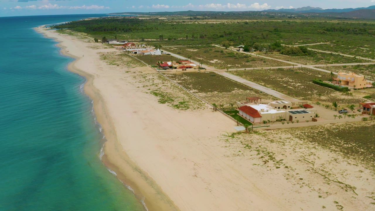 Diagonal aerial over La Ribera showing green-blue water, smooth sand, low surf, nearby houses and roads, inland vegetation, distant mountains, and no visible people.