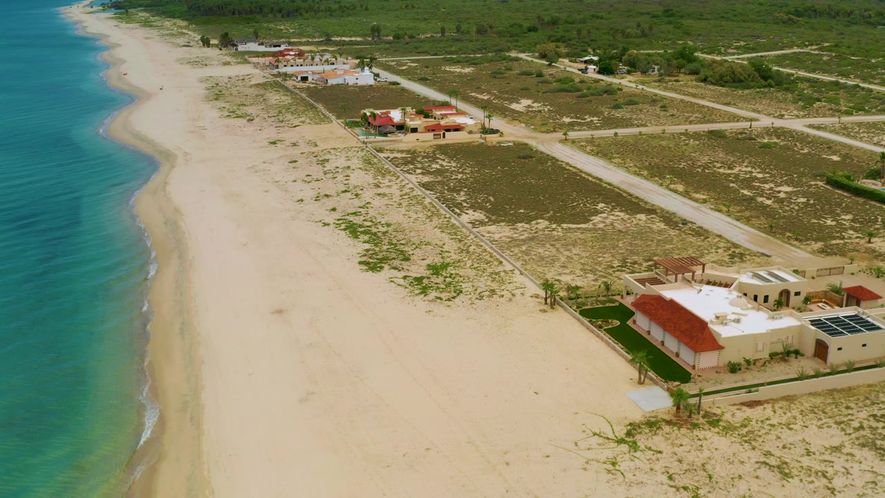 High aerial frame over La Ribera, Baja California Sur, with turquoise water on the left, a wide sandy beach, beachfront houses at the lower right, and no visible people.