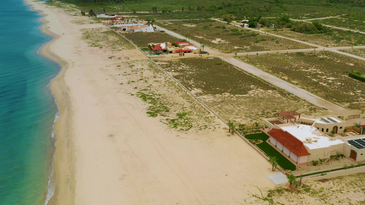 Aerial frame over La Ribera with the ocean tight to the left edge, broad empty sand below, a cluster of red-roof and white-roof homes to the right, and no visible people.