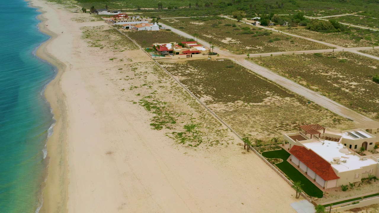 Aerial shot of sandy beach, surf line, houses, road, and scrubland.