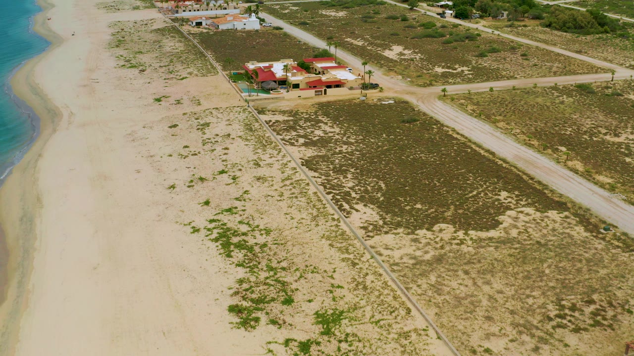 High aerial view in La Ribera, Baja California Sur with blue water along the left edge, pale sand, scrub-covered lots, dirt roads, and red-roofed buildings in the upper midground, with no visible people.