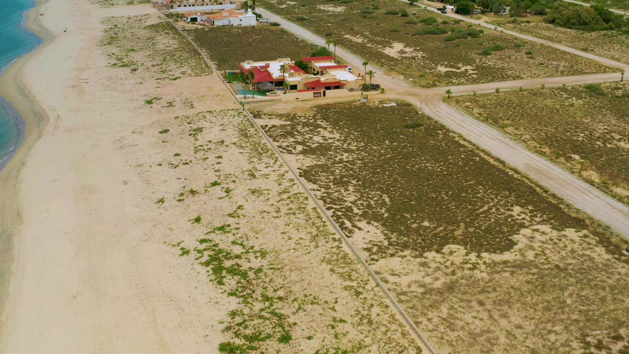 High oblique view over La Ribera, Baja California Sur with the beach and sea running down the left side, red-roofed buildings set back from the sand, and broad scrub lots divided by dirt roads, with no visible people.