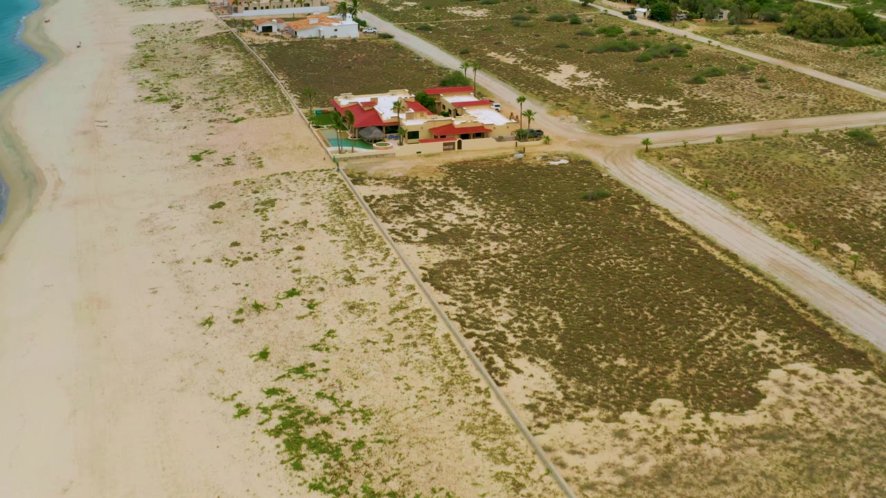 Aerial view of a red-roofed coastal property beside sand, scrubland, and dirt roads.