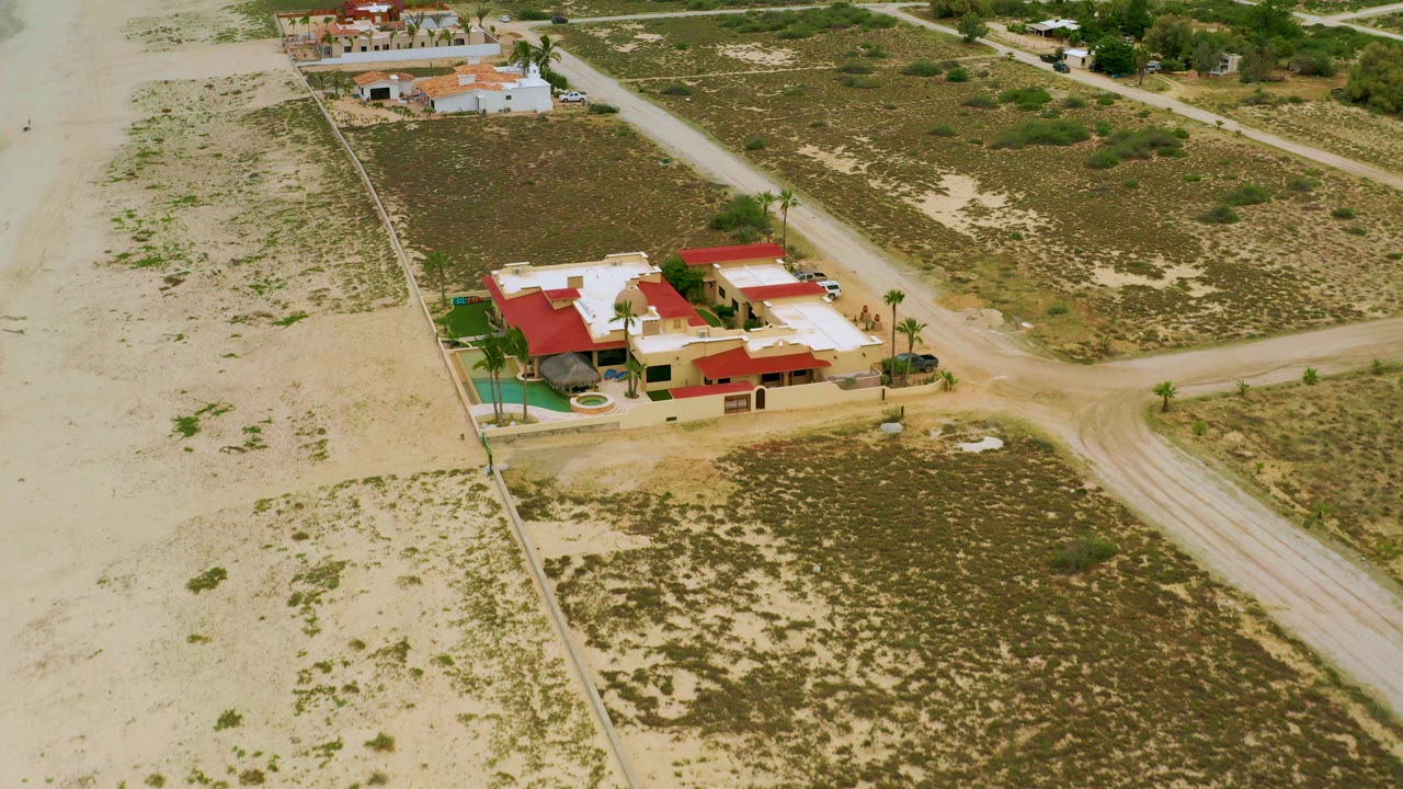 High aerial frame over a red-roof resort compound in La Ribera, Baja California Sur, with sandy lots, scrub vegetation, dirt roads, nearby homes, parked vehicles, and no visible people.