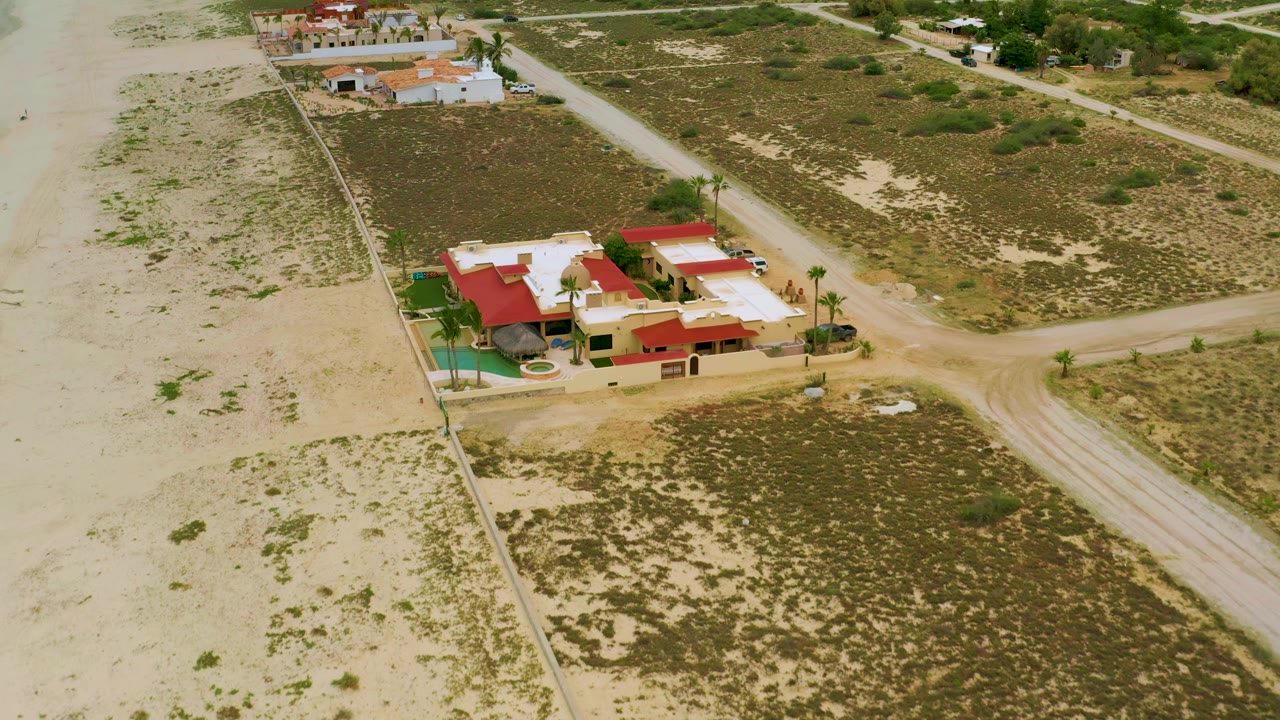 Aerial view in La Ribera, Baja California Sur, showing the resort slightly closer to center with red roofs, a pale roof deck, a turquoise pool area, dirt roads, desert scrub, and no visible people.