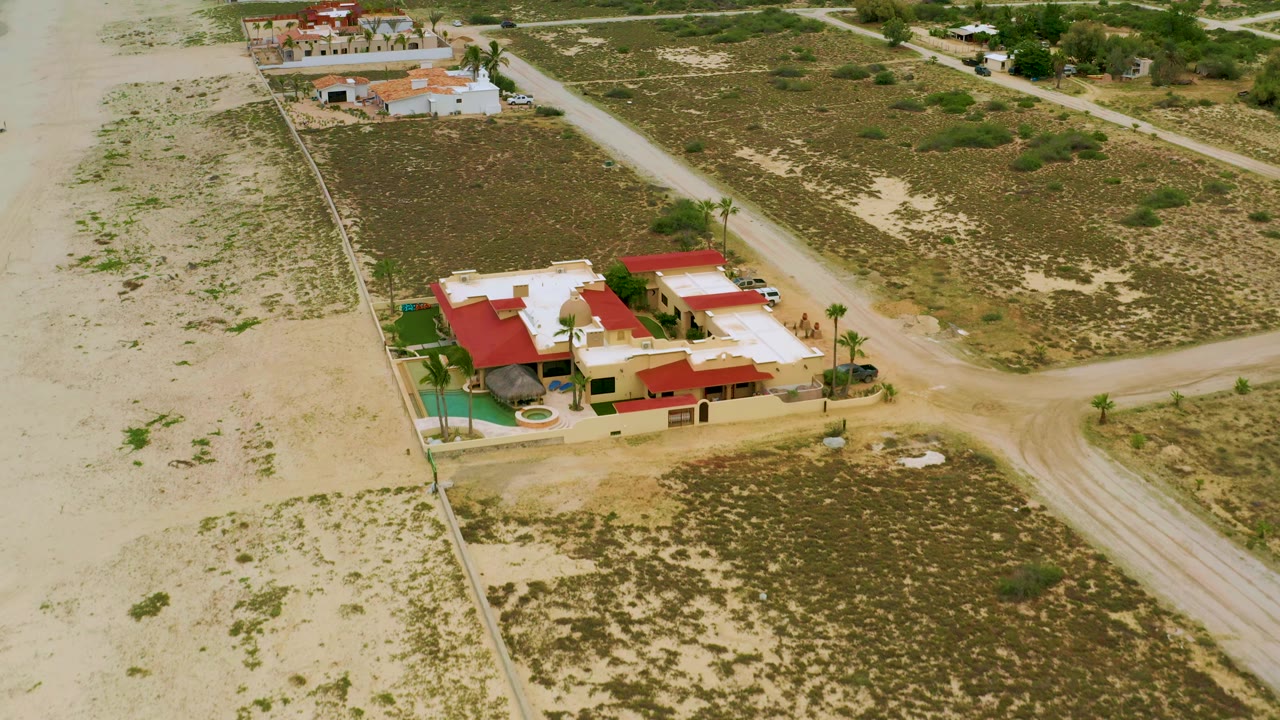 High drone-style frame over La Ribera, Baja California Sur, with the red-roof resort near the lower center, palms along the wall, sandy desert lots on both sides, dirt tracks, and no visible people.