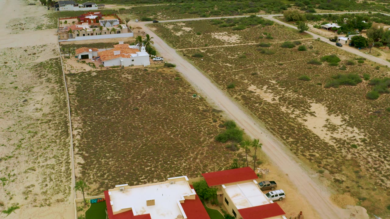 Aerial frame over La Ribera, Baja California Sur, showing a diagonal dirt road beside sandy scrub lots and white homes with red roofs, with no visible people.
