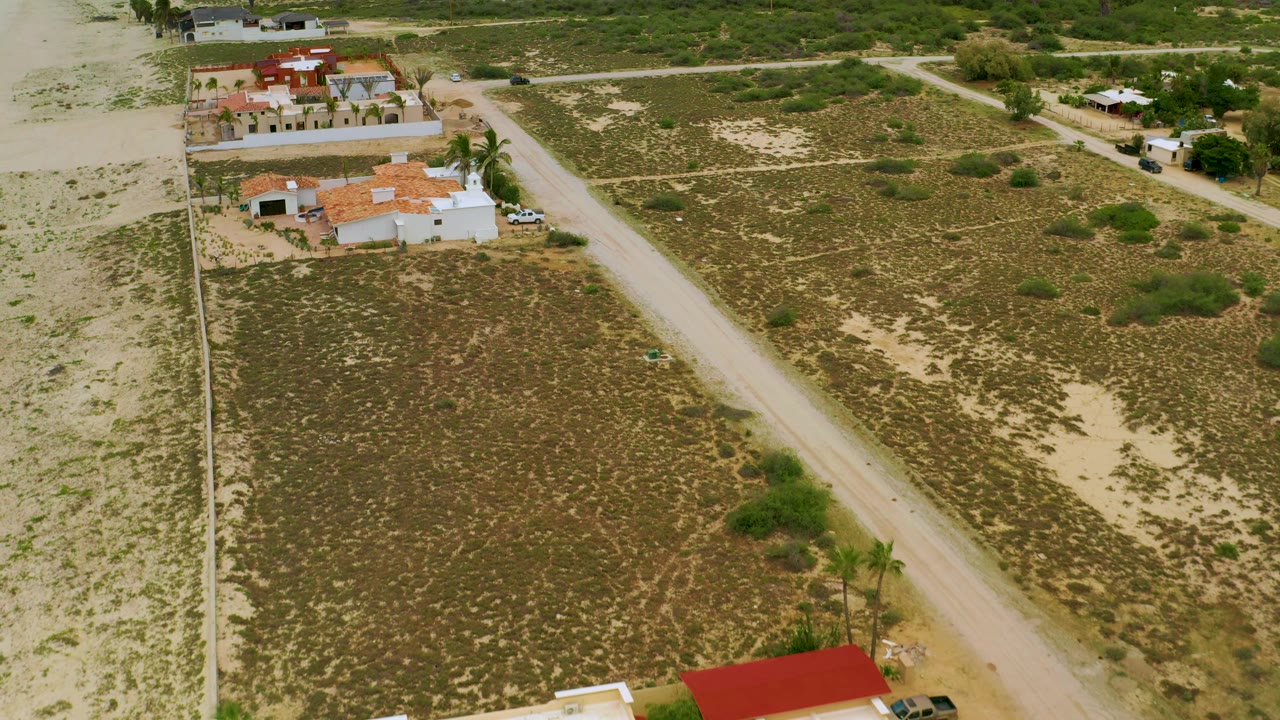 Aerial frame over La Ribera showing intersecting dirt roads, open sandy parcels, scattered green shrubs, and white residential buildings with no visible people.