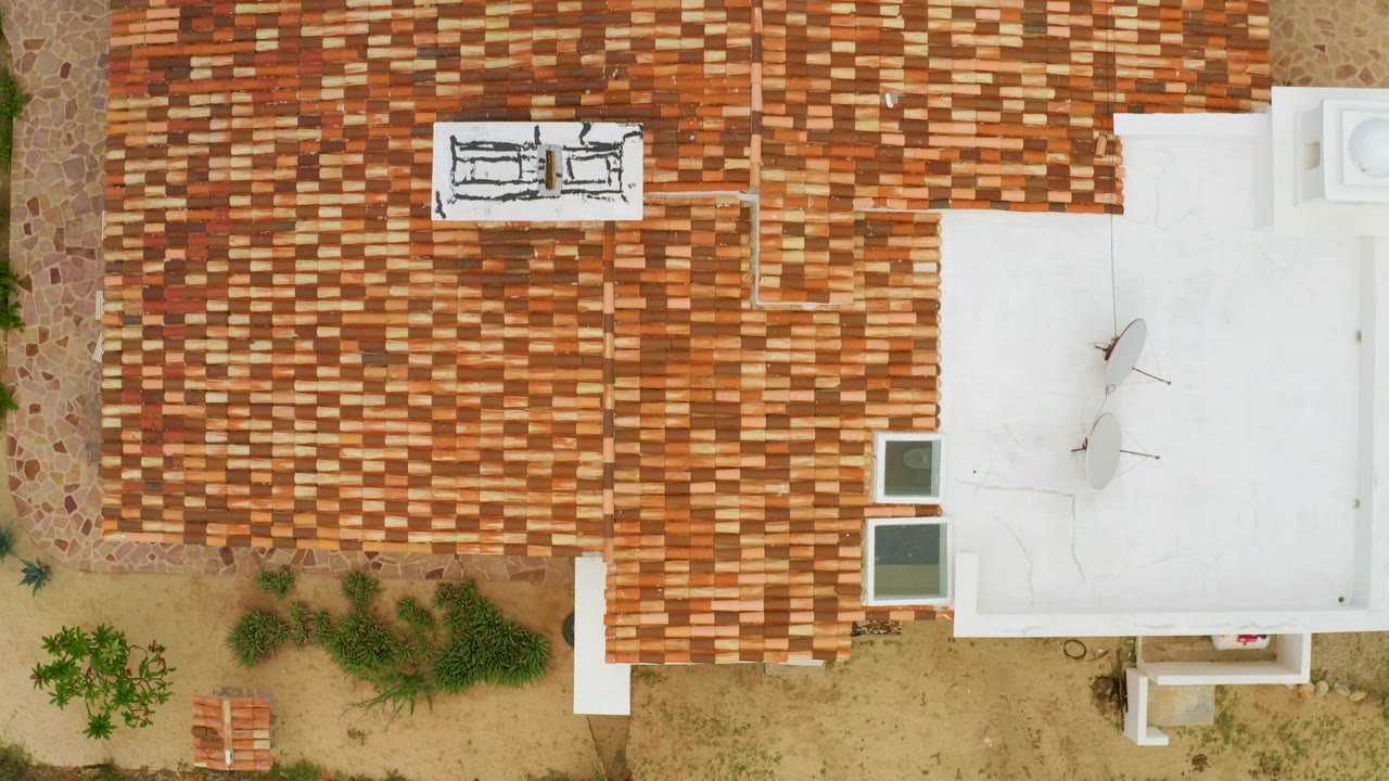 Top-down aerial frame in La Ribera, Baja California Sur showing a terracotta tiled roof beside a white flat roof, sandy garden ground, scattered shrubs, and no visible people.