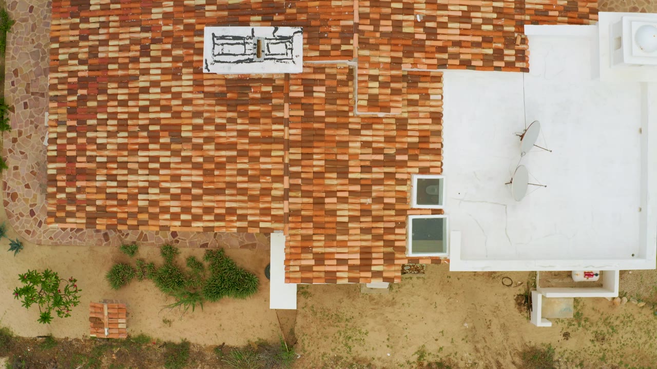 Top-down La Ribera frame showing more of the paved driveway edge at left, the tiled roof across the upper area, the white roof at right, sandy garden below, and no visible people.