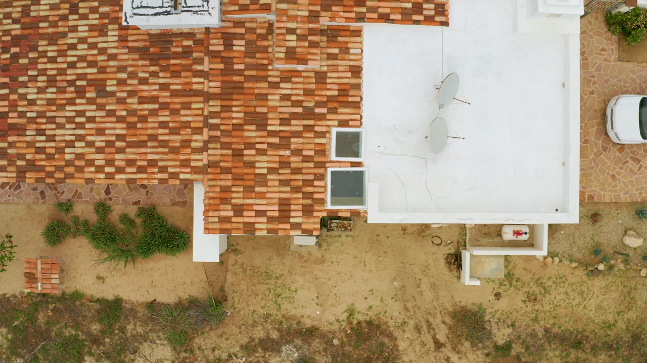 Top-down aerial frame over La Ribera showing red tile roofs, a flat white roof, sandy ground, sparse shrubs, and a white vehicle near the upper right with no visible people.