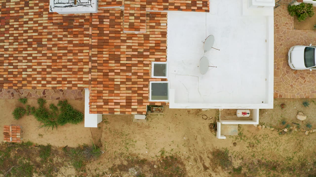 Aerial frame over La Ribera showing patterned clay roof tiles, a flat white roof, dry soil, planted shrubs, and a partial white vehicle at the top edge with no visible people.