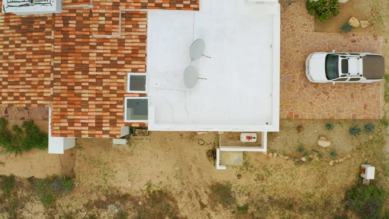 Overhead frame in La Ribera, Baja California Sur showing a red tile roof, flat white roof, parked white car, sandy garden ground, stone paving, and no visible people.