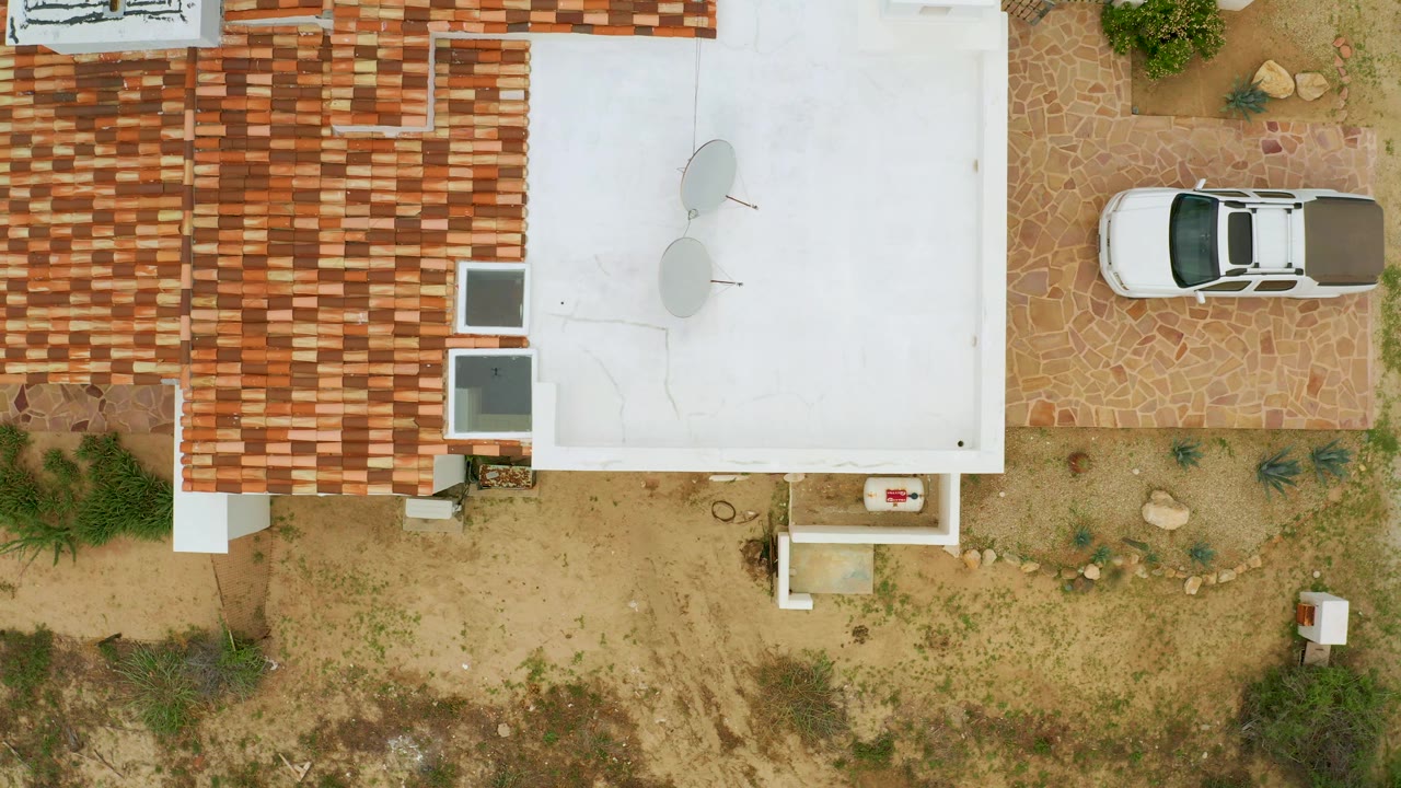 Top-down La Ribera frame with the parked white car near the upper left, patterned red roof tiles across the center, a white roof section, sparse plants, and no visible people.