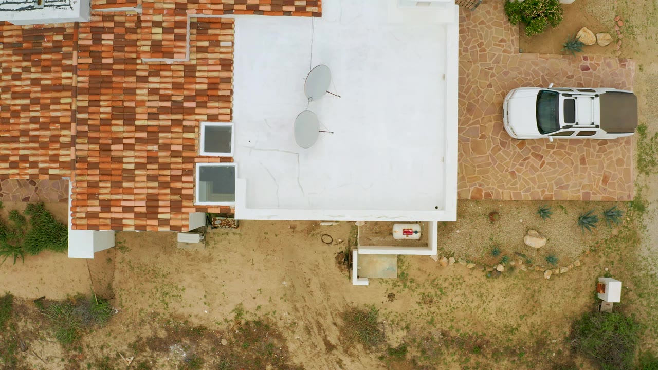 Aerial frame in La Ribera showing the white car on stone paving, red and tan roof tiles, a bright white roof with two dishes, dry landscaping, and no visible people.