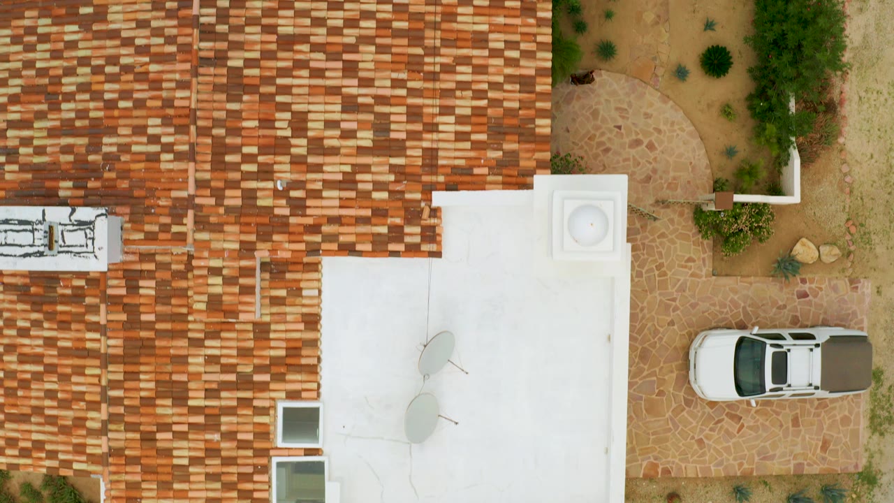 Overhead aerial frame in La Ribera, Baja California Sur, Mexico, showing patterned tile roofs, a white building section, sandy courtyard planting, a parked white car, and no visible people.