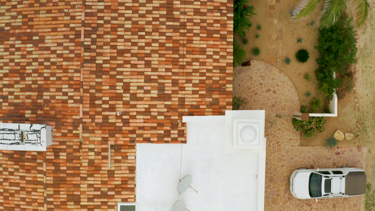 Aerial frame in La Ribera, Baja California Sur, Mexico, showing a dense field of roof tiles, a white rooftop area, sandy courtyard vegetation, a partly framed parked white car, and no visible people.