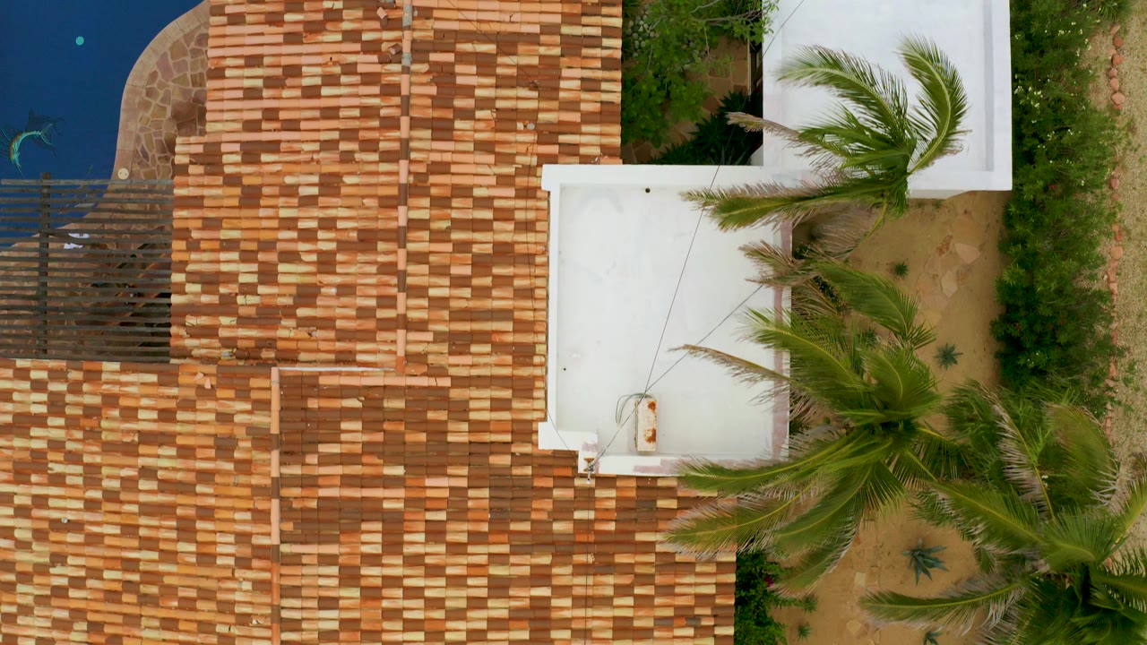 Top-down aerial frame in La Ribera, Baja California Sur, showing a terracotta tile-roofed residence beside a blue pool, palms, sandy yard, and white roof structures, with no visible people.