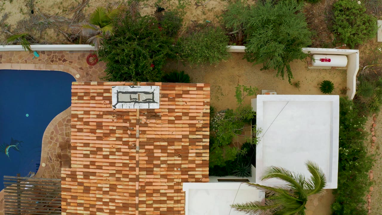 Second overhead frame in La Ribera shows the blue pool partly at left, terracotta roof centered, white roofs at right, garden shrubs, sandy ground, and no visible people.