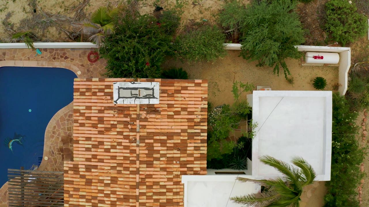 Overhead La Ribera frame with more of the pool visible at left, a large terracotta tiled roof across the center, white building edges, green trees, sand, and no visible people.