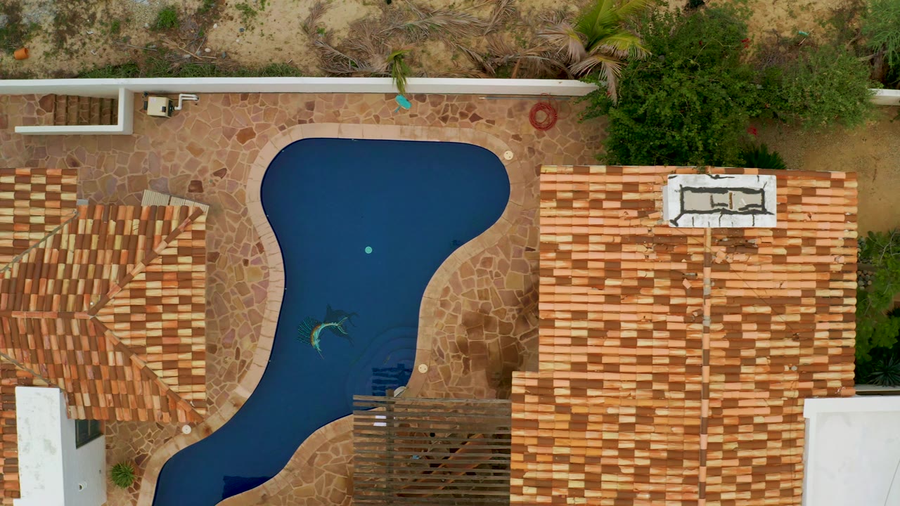 Bird's-eye La Ribera frame with the pool slightly left of center, terracotta and tan shingle roofs on both sides, patio stonework, and no visible people.