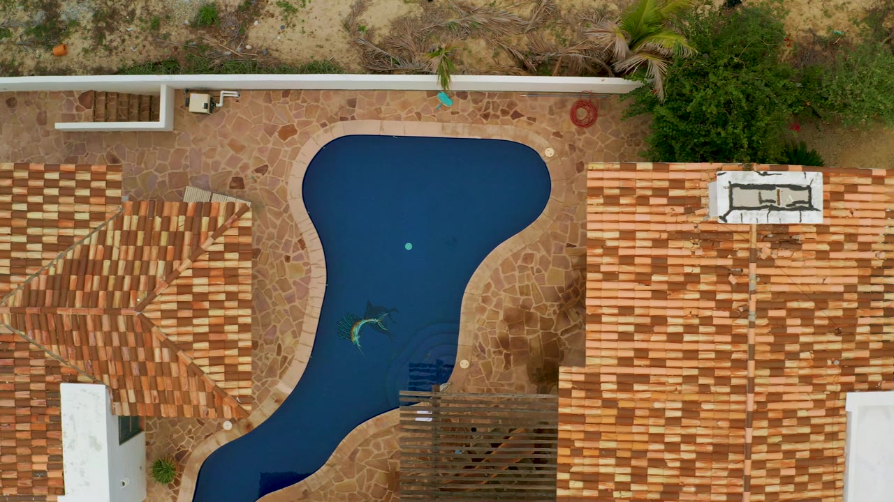 Top-down La Ribera frame showing the dark pool, patterned roofs, tan paving, dry soil beyond the wall, and no visible people in the residential scene.