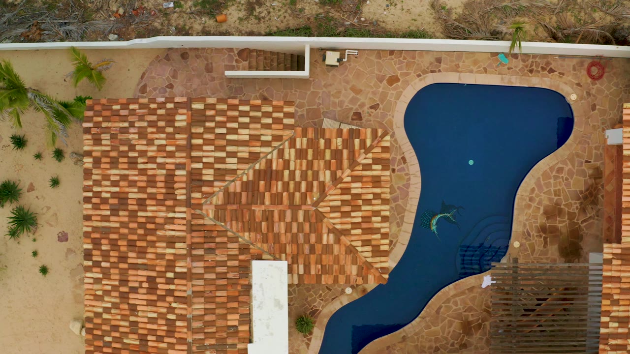 Vertical aerial frame in La Ribera, Baja California Sur centered on the patterned tile roof beside a bean-shaped dark pool, with stone paving, sparse shrubs, and no visible people.