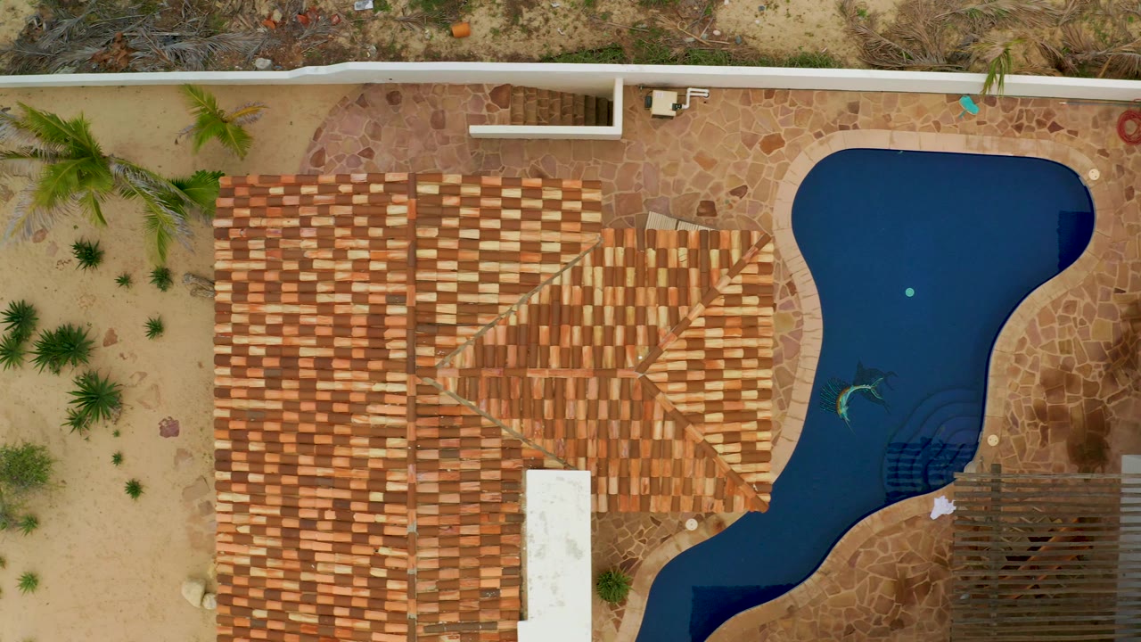 Overhead La Ribera frame with the tiled roof occupying the center, the pool along the right edge, sandy landscaping and palms to the left, and no visible people.