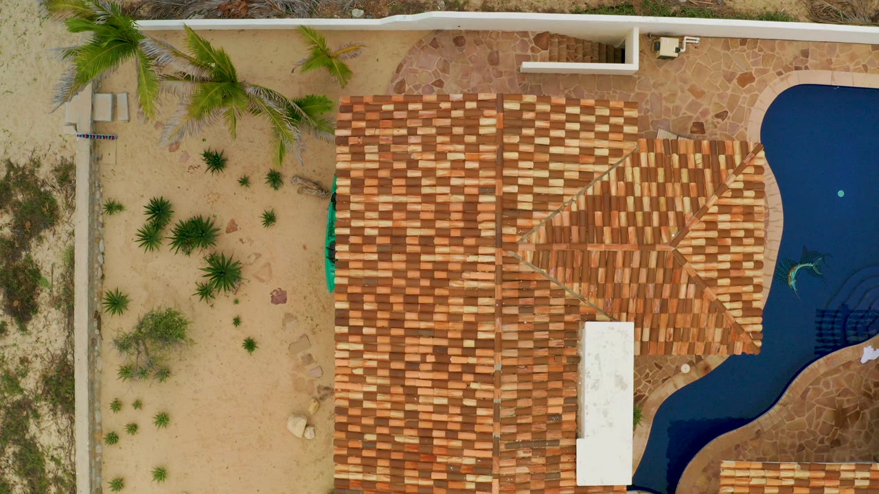 Straight-down aerial frame in La Ribera, Baja California Sur, showing terracotta tile roofs, tan sand, palms, stone paths, and a curved blue pool edge with no visible people.