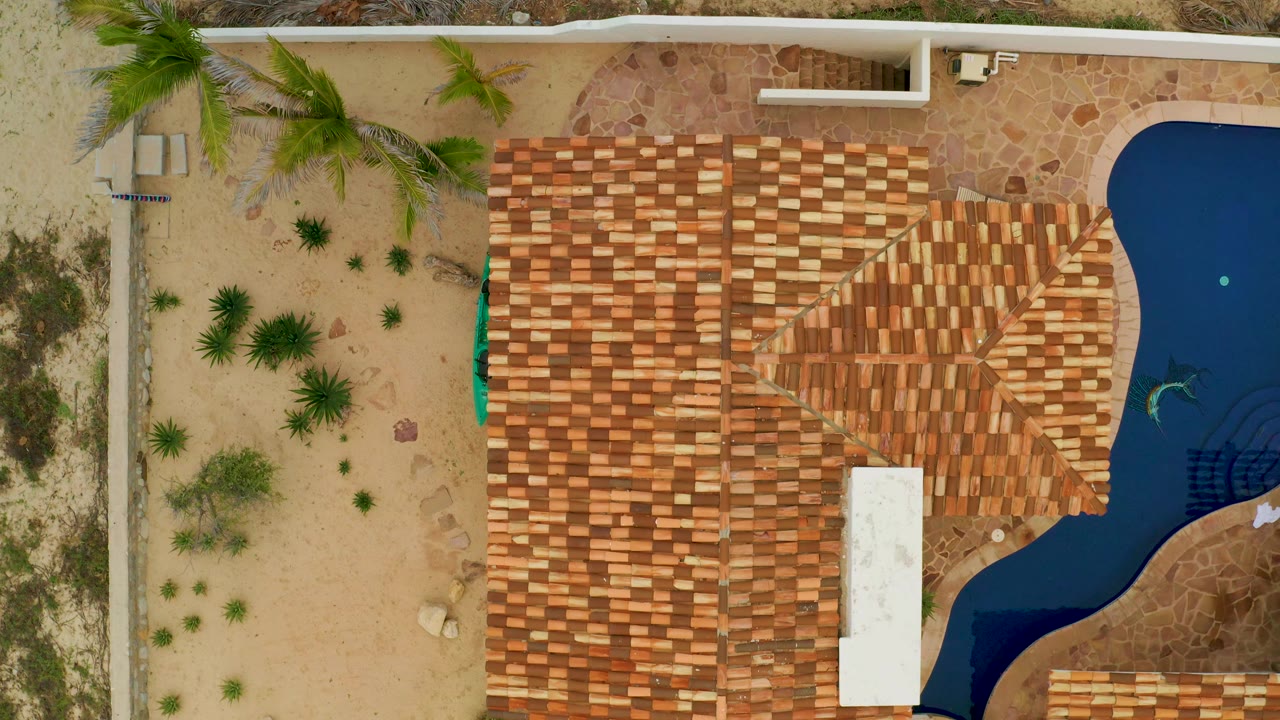 Overhead La Ribera frame with a broad tiled roof mass centered, sparse desert plants at left, and dark blue pool water along the right side, with no visible people.