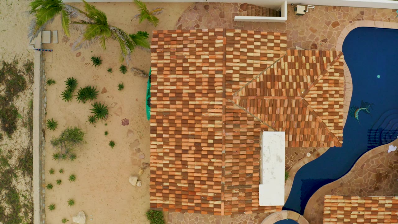 Aerial La Ribera view showing the roof shifted farther right, a sandy landscaped strip on the left, a white rectangular structure on the roof area, and pool water at the right edge with no visible people.