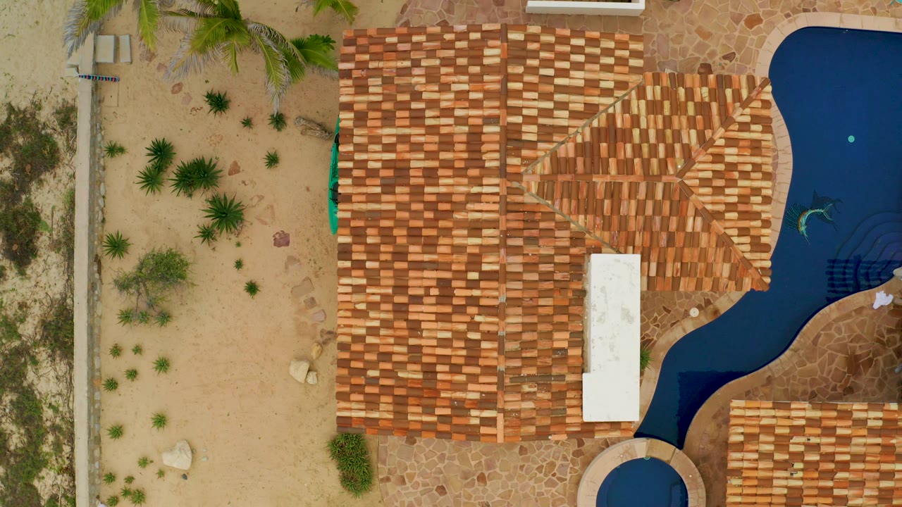 Tile roof complex beside blue pool water and a sandy planted area.