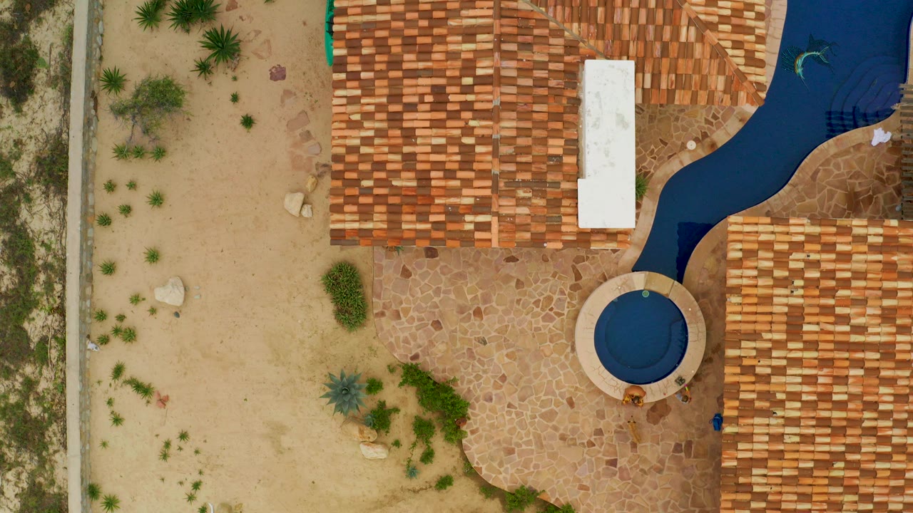 Top-down aerial frame in La Ribera, Baja California Sur, showing tan sand at left, terracotta roof sections, stone paving, a curved dark blue water feature, and a round blue pool with no visible people.