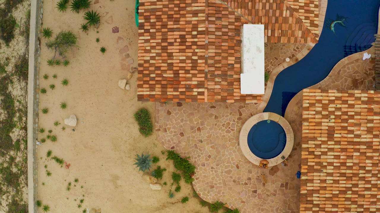 Second overhead frame in La Ribera shows the round blue pool near the right side, terracotta tiled roofs above and below it, tan sand and scattered plants to the left, and no visible people.
