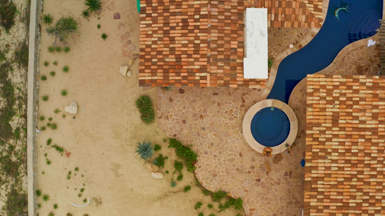 Aerial frame in La Ribera with more sandy open ground visible on the left, a circular blue pool near the tiled roof edge, stone paving around it, and no visible people.