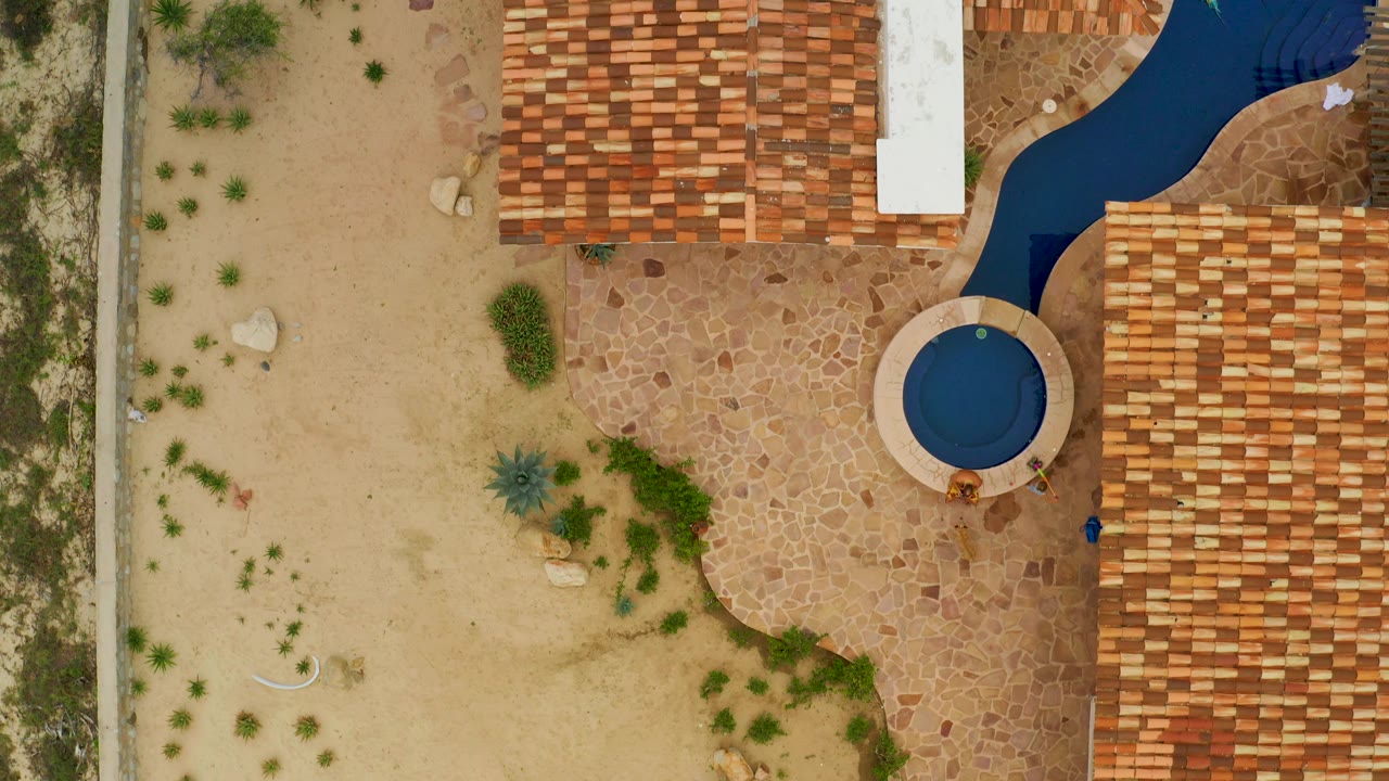 Drone-like overhead view of sand, shrubs, roof tiles, paving, and a small round pool.